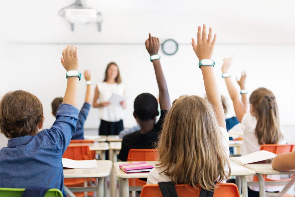 Classroom scene with students raising hands, teacher at the front.